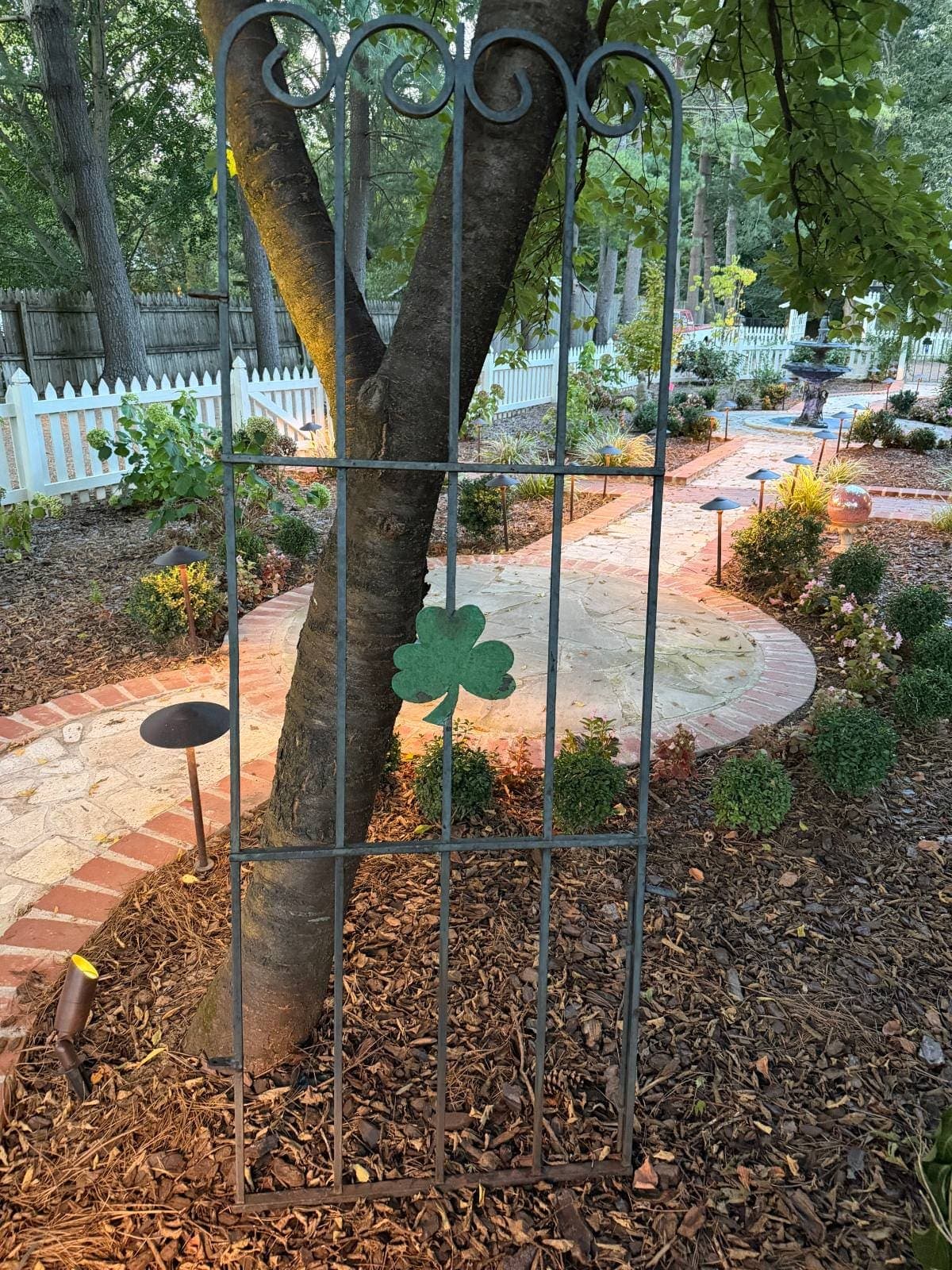 Antique iron gate with scrollwork and a green shamrock, leaning against a tree before restoration
